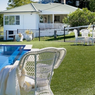 A private holiday home with a green lawn and an outdoor pool surrounded by scattered seating and a black bar fence. There is blue sky and mountains in the distance.