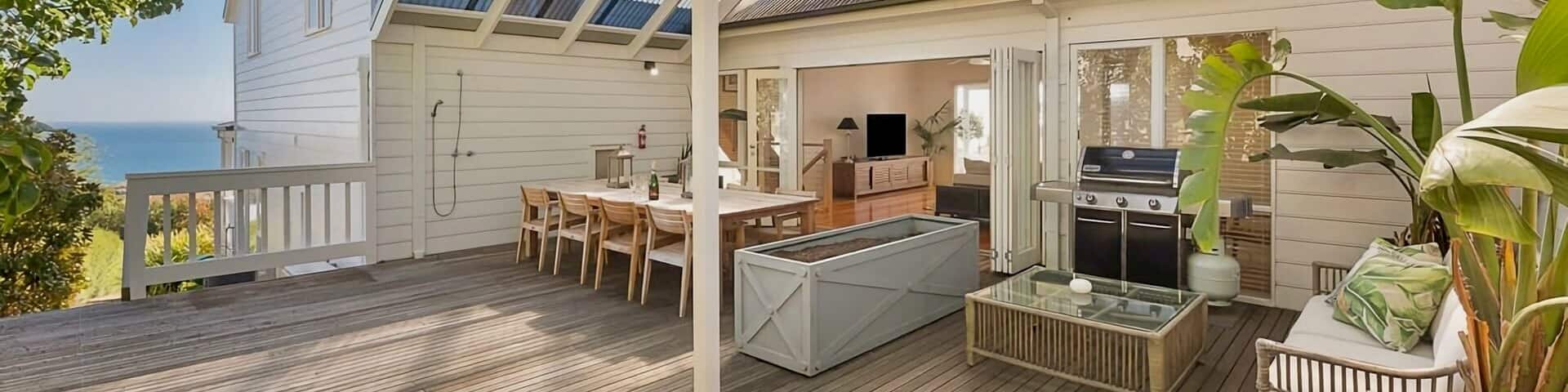 A shaded terrace with a large dining table, BBQ and soft seating area. Open patio doors show a lounge with a widescreen TV.
