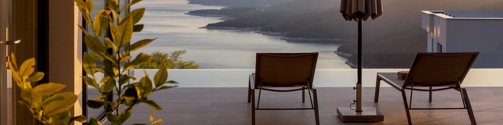 Une terrasse avec des chaises et un parasol donne sur une piscine Ă dĂ©bordement face Ă lâocĂ©an et aux collines verdoyantes. Le soleil se montre sur les collines.