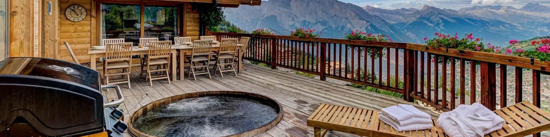 Sprudelnder, in den Boden eingelassener Whirlpool inmitten einer großen Terrasse eines wunderschönen Holzchalets mit herrlicher Aussicht auf die Berge.