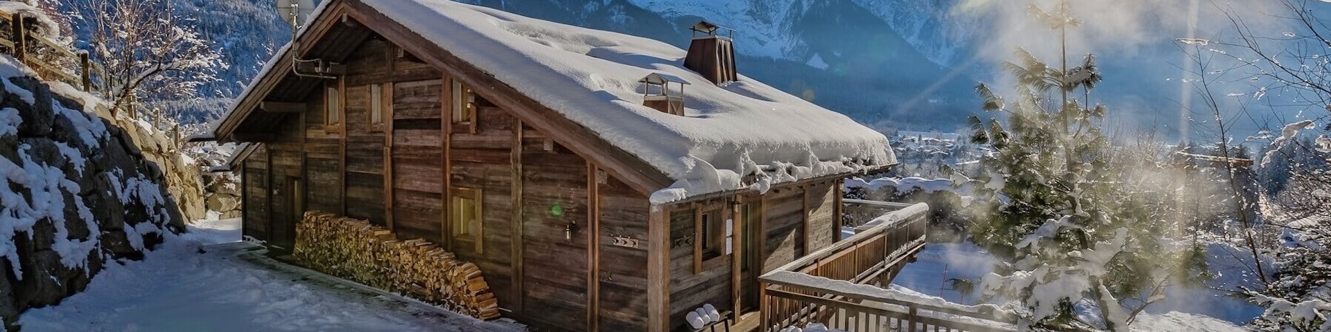 Un chalet de ski en bois au toit enneigé se dresse au milieu d’arbres clairsemés et face à des montagnes enneigées.