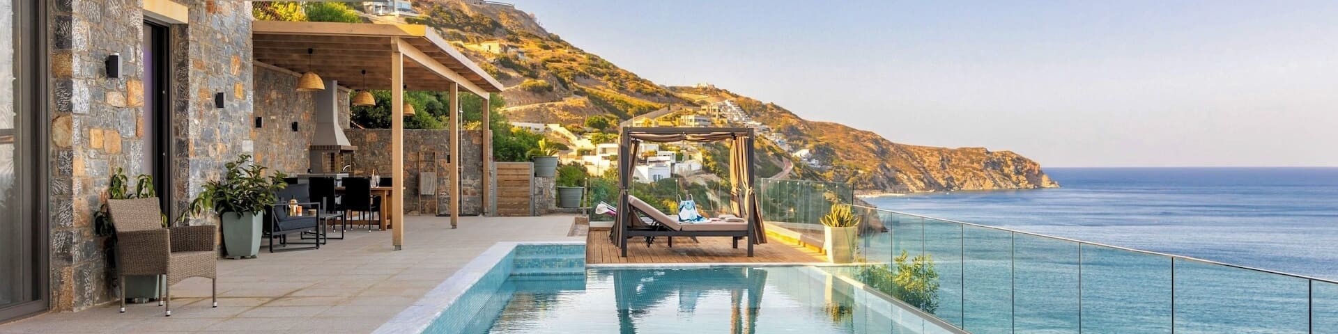Une terrasse près de l’eau donnant sur la mer et les montagnes, avec une piscine, des transats et un coin repas sous pergola.