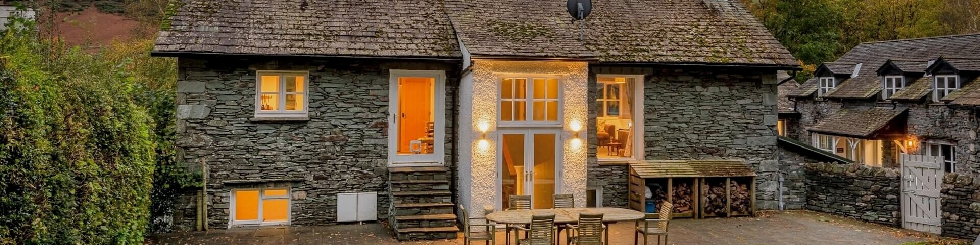 A cosy stone cottage at dusk surrounded by trees, with light shining through the windows.