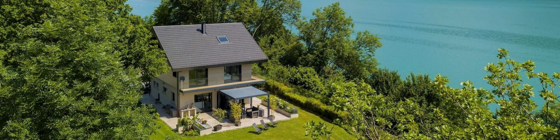 Ein zweistöckiges Haus umgeben von grünem Rasen und Bäumen. Im Hintergrund befinden sich ein See und bewaldete Hügel.