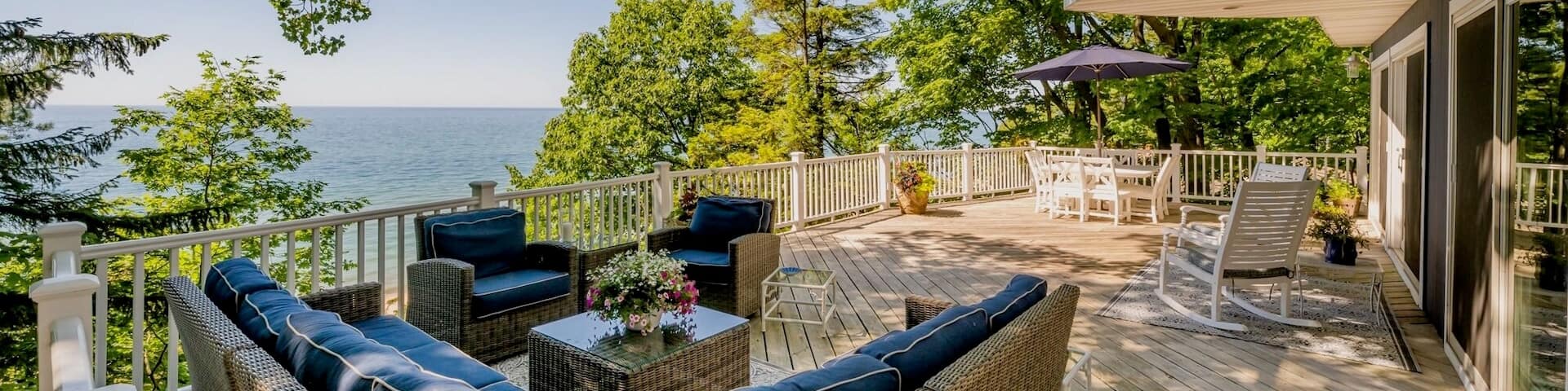 Un canapé d’extérieur en demi-cercle fait face à une table, à d’autres chaises et à l’océan depuis un balcon en bois bordé de branches d’arbres.