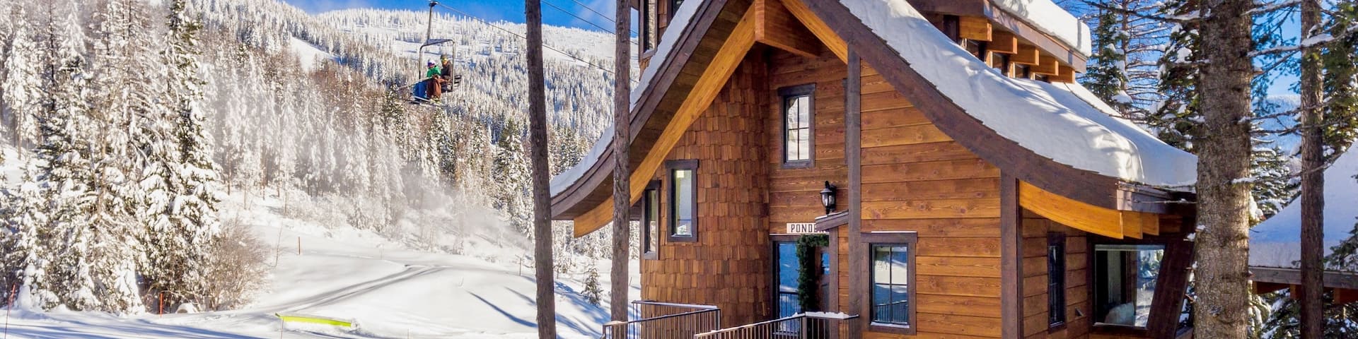Un chalet de ski en bois au toit couvert de neige se dresse devant une forĂȘt enneigĂ©e. DerriĂšre le chalet, deux personnes montent en tĂ©lĂ©siĂšge.