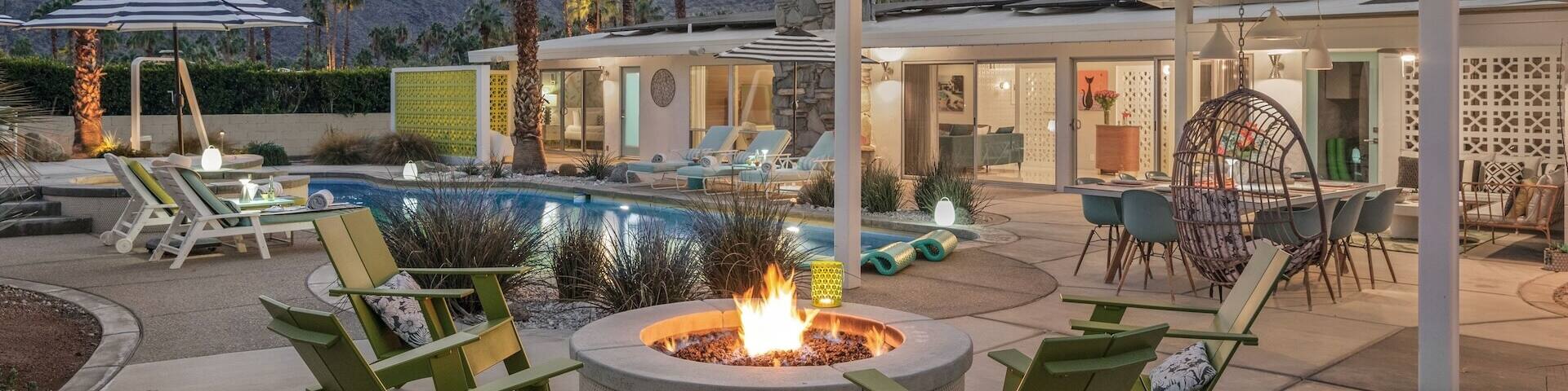 A large property at dusk with an outdoor pool, seating areas and a lit firepit with mountains in the background.