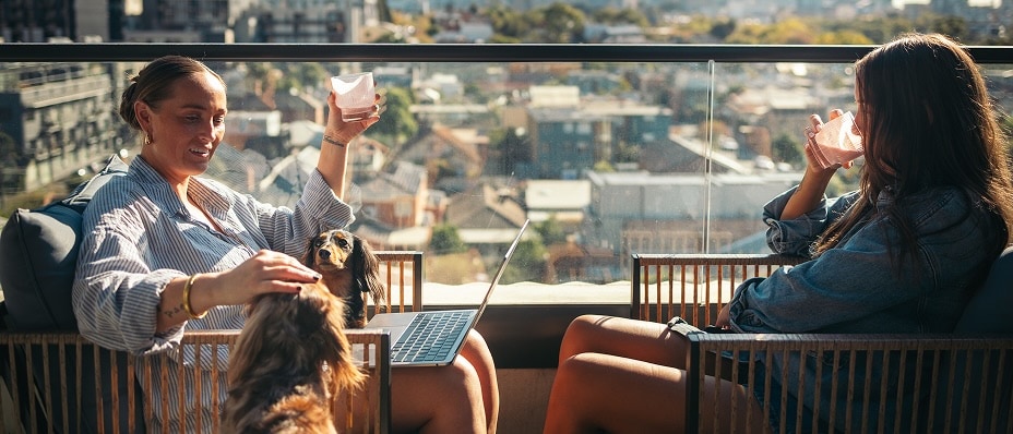 Two women sit on a balcony with drinks and a dog, overlooking a sunlit cityscape.