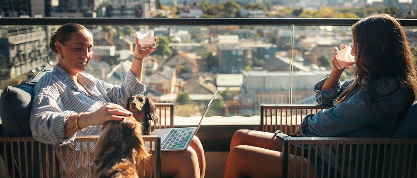 Two women sit on a balcony with drinks and a dog, overlooking a sunlit cityscape.