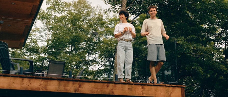 Deux personnes sont debout sur une terrasse en bois surélevée, à côté d’une maison moderne, entourée de grands arbres.