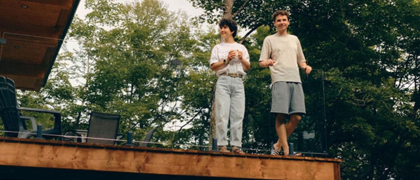 Deux personnes sont debout sur une terrasse en bois surélevée, à côté d’une maison moderne, entourée de grands arbres.