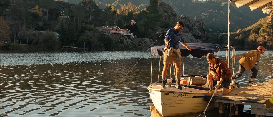 Three men on a wooden dock on a lake, surrounded by a hilly, wooded landscape on a sunny day.