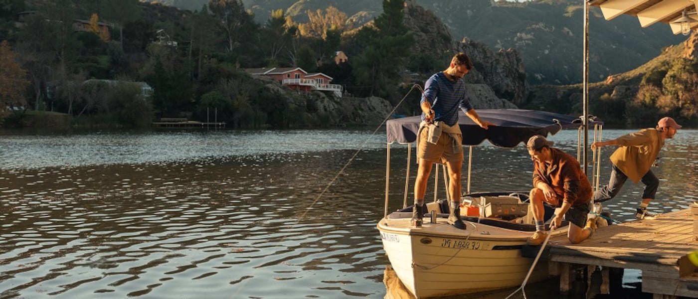 Three men on a wooden dock on a lake, surrounded by a hilly, wooded landscape on a sunny day.