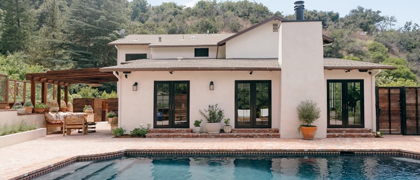 The rear exterior of a Mediterranean-style house with a brick patio, a swimming pool and a hillside in the background.