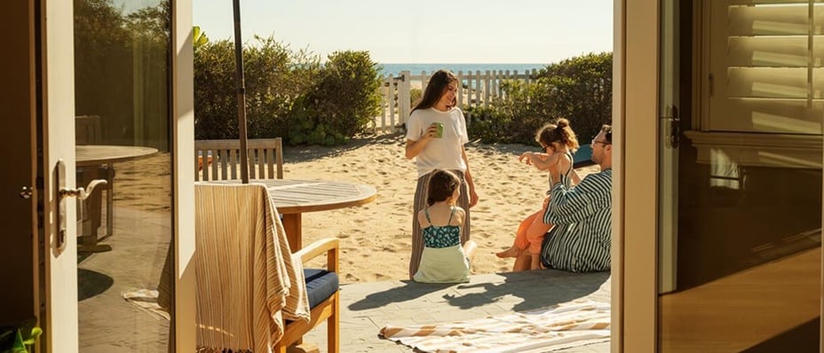 A family relaxing on a sandy patio bordered by a white picket fence that leads directly onto a beach.