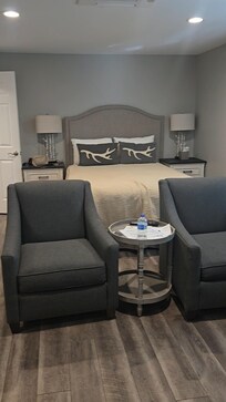 Bathroom to the left, full-size bed, two chairs, small table. Big screen tv mounted on the wall to the right above a fireplace.