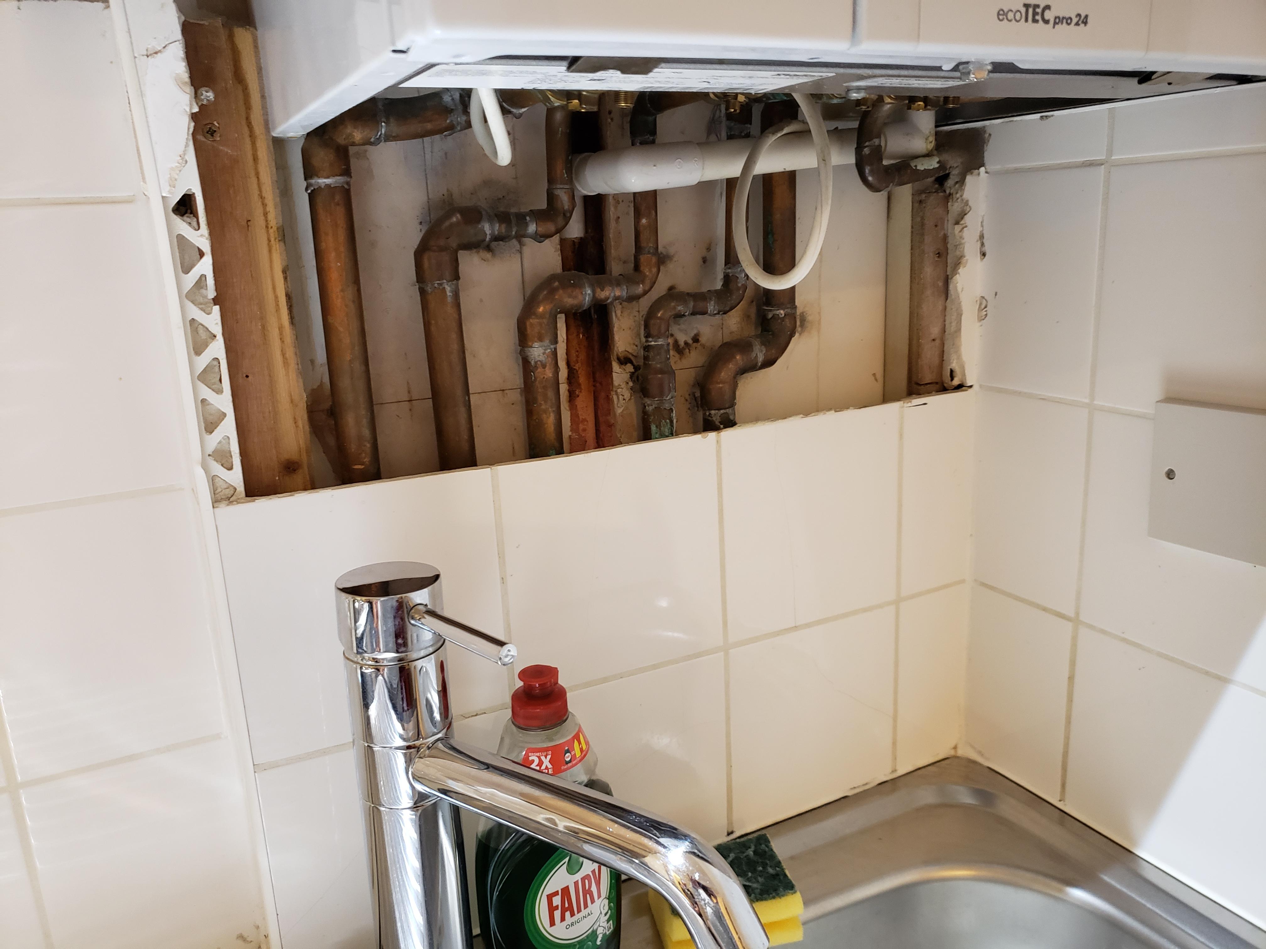 Broken tiles and exposed pipes on kitchen walls