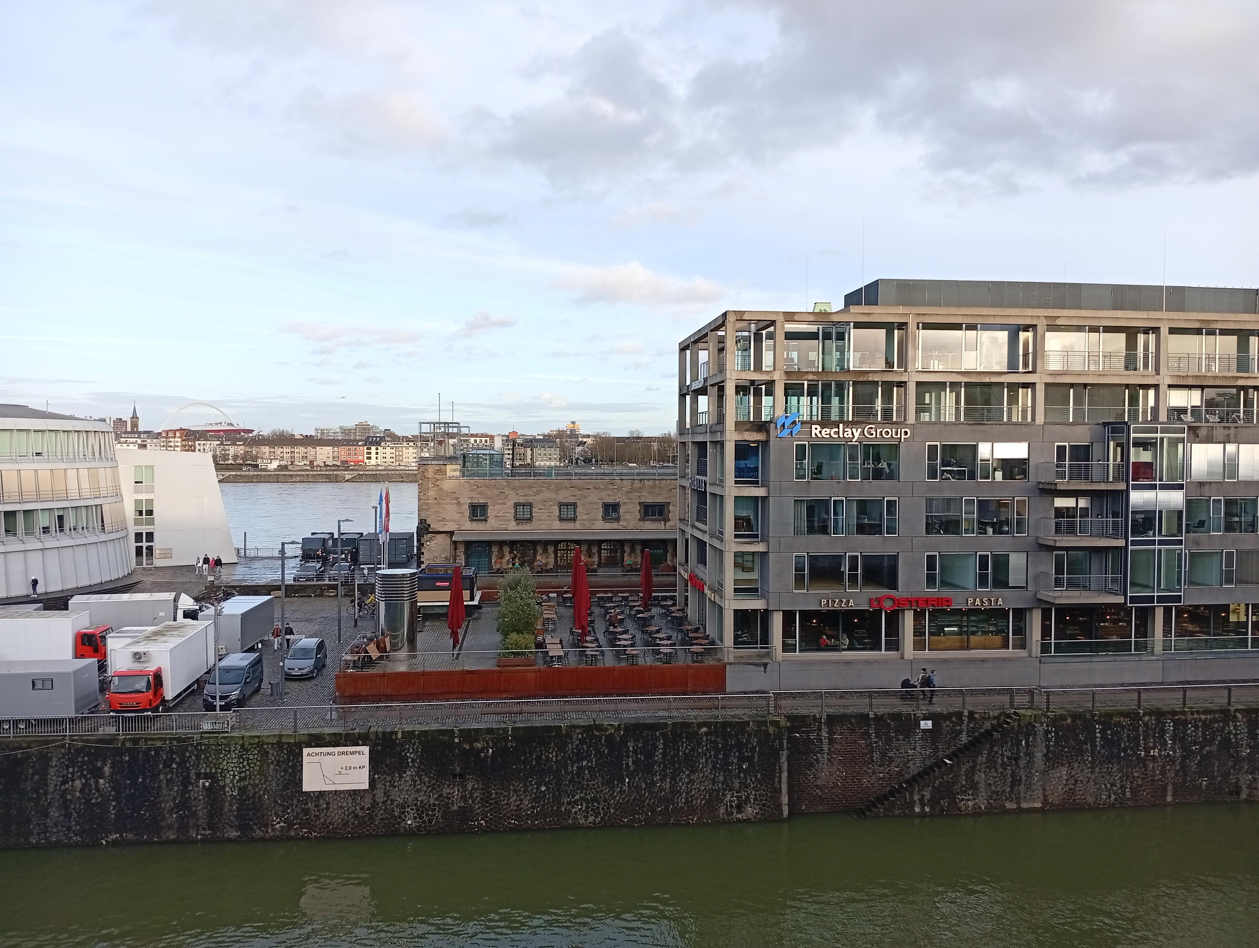 Blick auf das gegenüber liegende Bürogebäude, dahinter der Rhein, davor ein Seitenarm vom Fluss