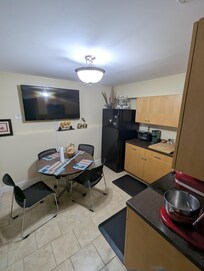 Dining room area, with cozy chairs and table, refrigerator, sink, TV, nicer placemats than the previous picture, etc. as of May 16, 2026. Not pictured: drawers containing forks, spoons, sharp knives, bowls, oven, stovetops, etc.,as are standard