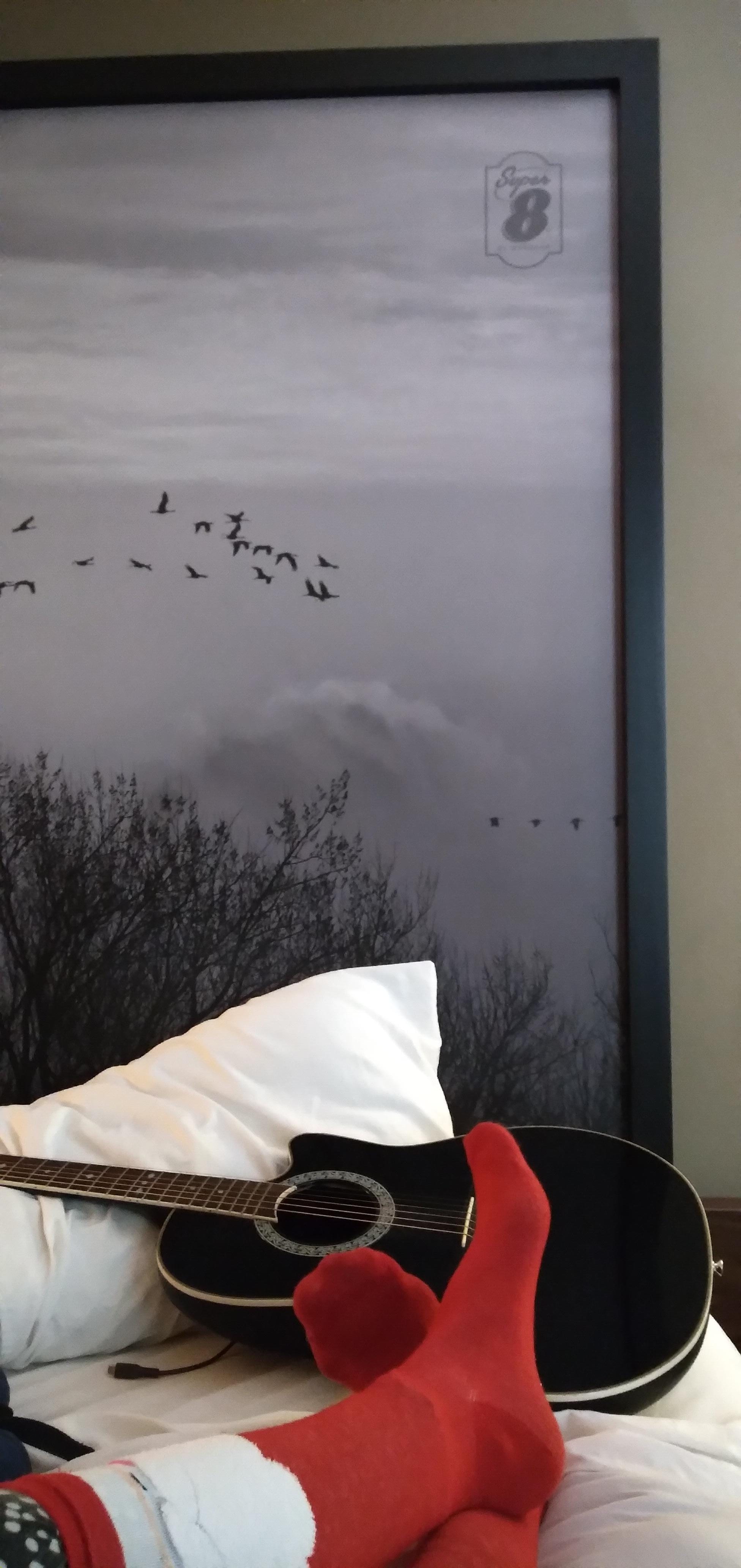 Photo of feet crossed wearing red socks, on a bed. Behind the feet there is a black acoustic guitar, and behind that a grey and black sky painting with black flying birds.