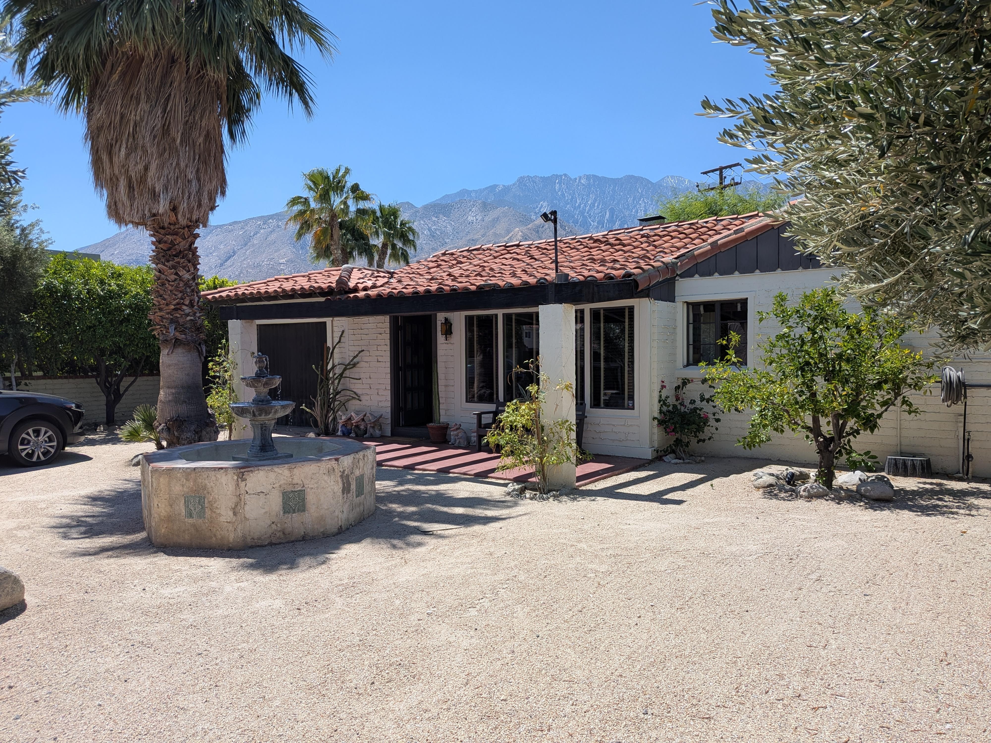 A bright, daytime shot of the front of the villa under a clear blue sky. It shows the red-tiled roof, the white brick exterior, and the large stone tiered fountain in the foreground. In the distance, the rugged San Jacinto Mountains are visible.