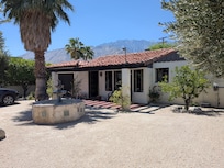 A bright, daytime shot of the front of the villa under a clear blue sky. It shows the red-tiled roof, the white brick exterior, and the large stone tiered fountain in the foreground. In the distance, the rugged San Jacinto Mountains are visible.
