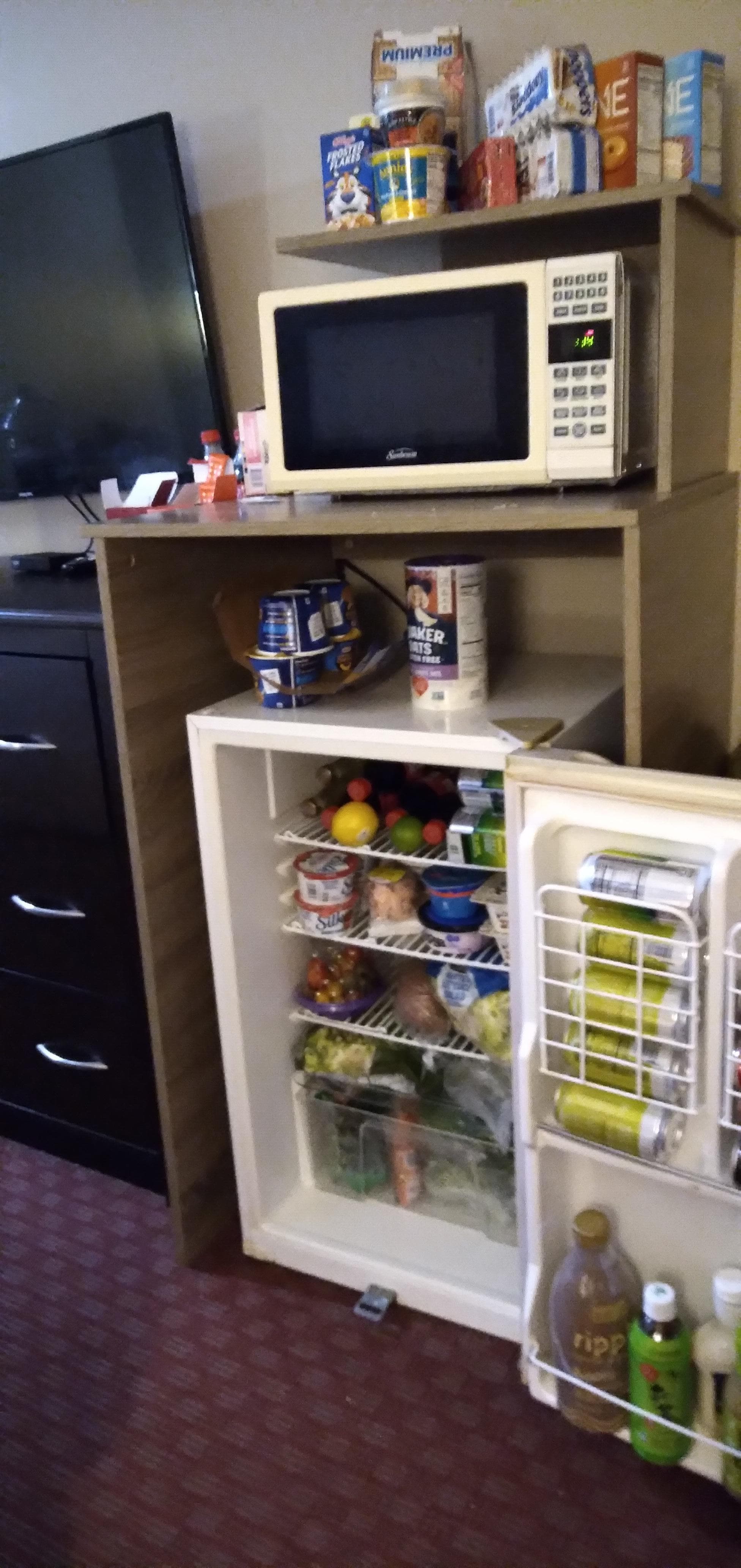 Photo of kitchenette area, fairly large mini fridge stocked full of salads, veggies, drinks, yogurt, milk, dressings. A shelf above the fridge holds a microwave. To the left of the fridge is a dresser. Above the dresser is a flat screen tv.
