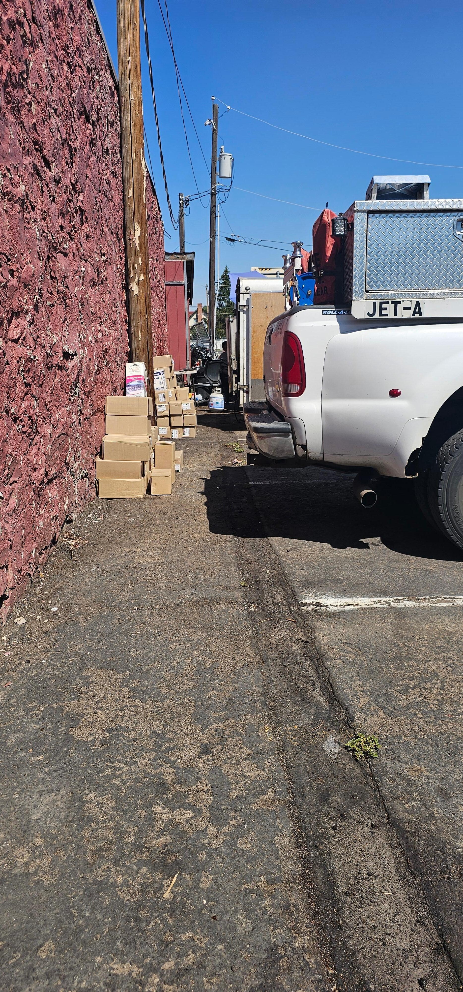 Boxes and junk stacked against building in parking lot.  People drinking and barbecuing between vehicles