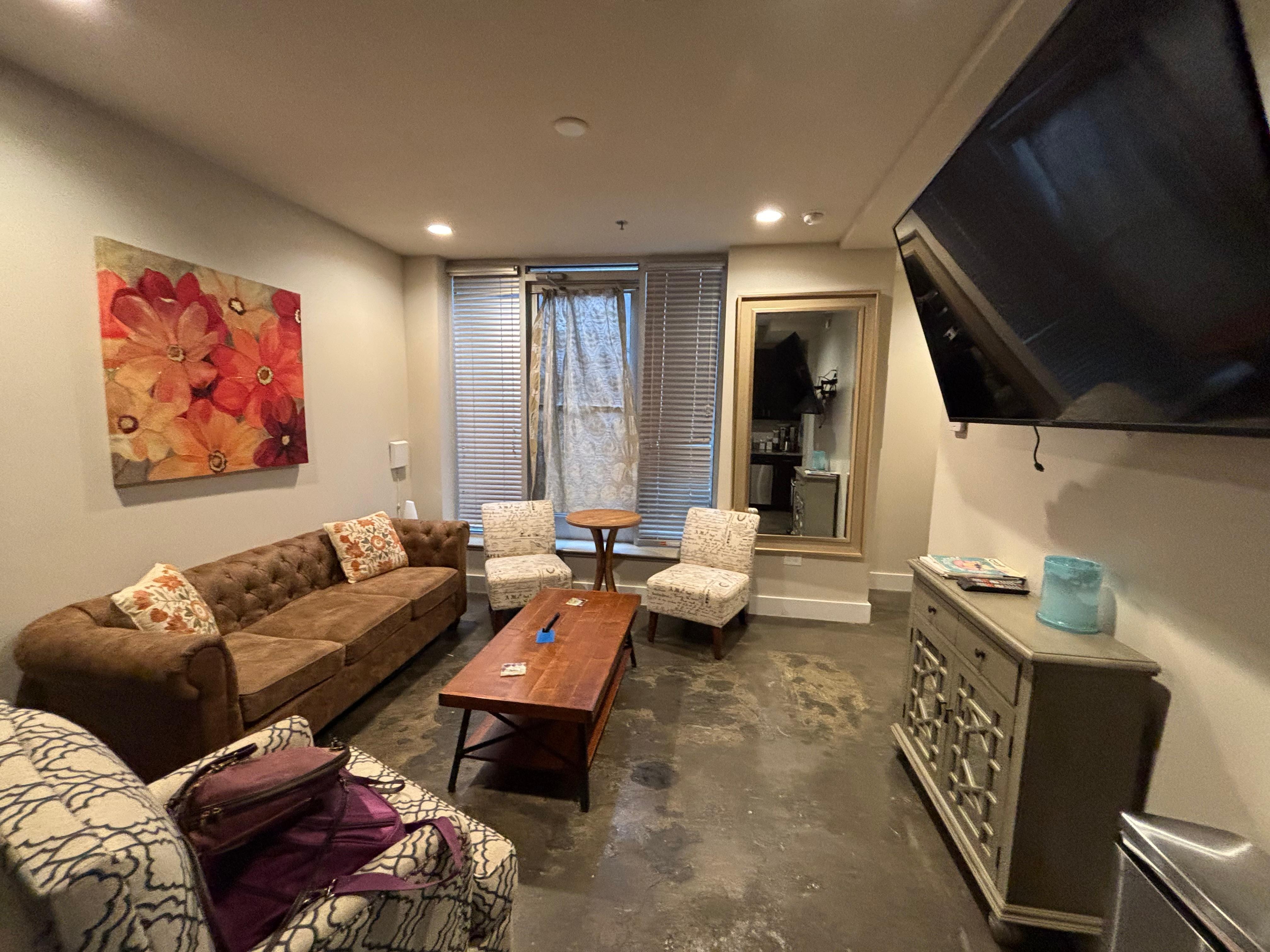 View of living room from kitchen. Full kitchen has sink, oven, microwave & Fridge. There’s a giant TV on the wall. 