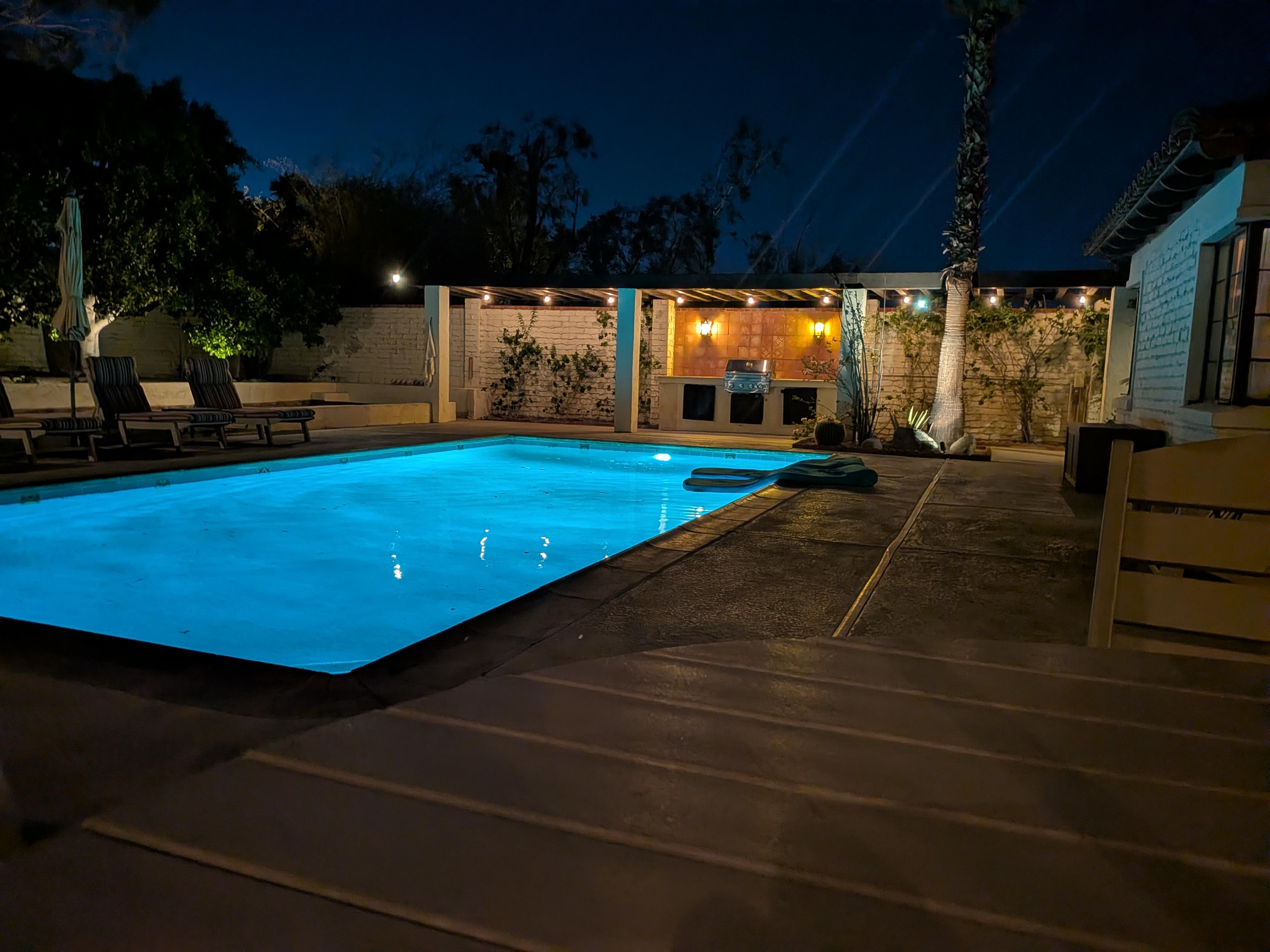 A dramatic nighttime shot of the backyard. The saline pool is glowing with bright blue light, contrasting against the dark sky. In the background, you can see the built-in  BBQ area under a pergola with warm string lights.