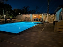 A dramatic nighttime shot of the backyard. The saline pool is glowing with bright blue light, contrasting against the dark sky. In the background, you can see the built-in BBQ area under a pergola with warm string lights.