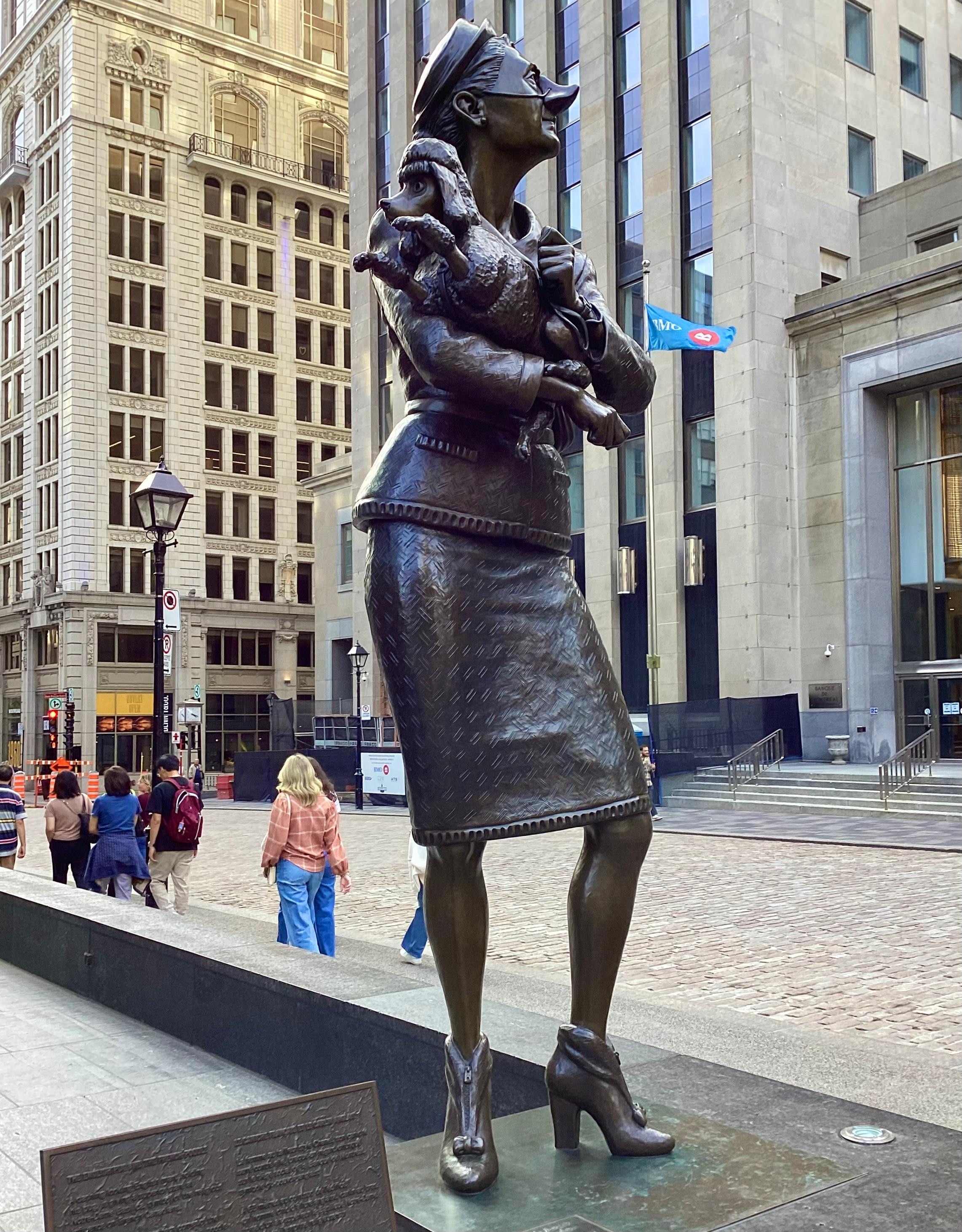 The haughy female statue n a Chanel-like suit, holding her poodle, the woman turning up her long, snooty nose at the nearby bank; the female statue is located on the corner opposite the snooty man.