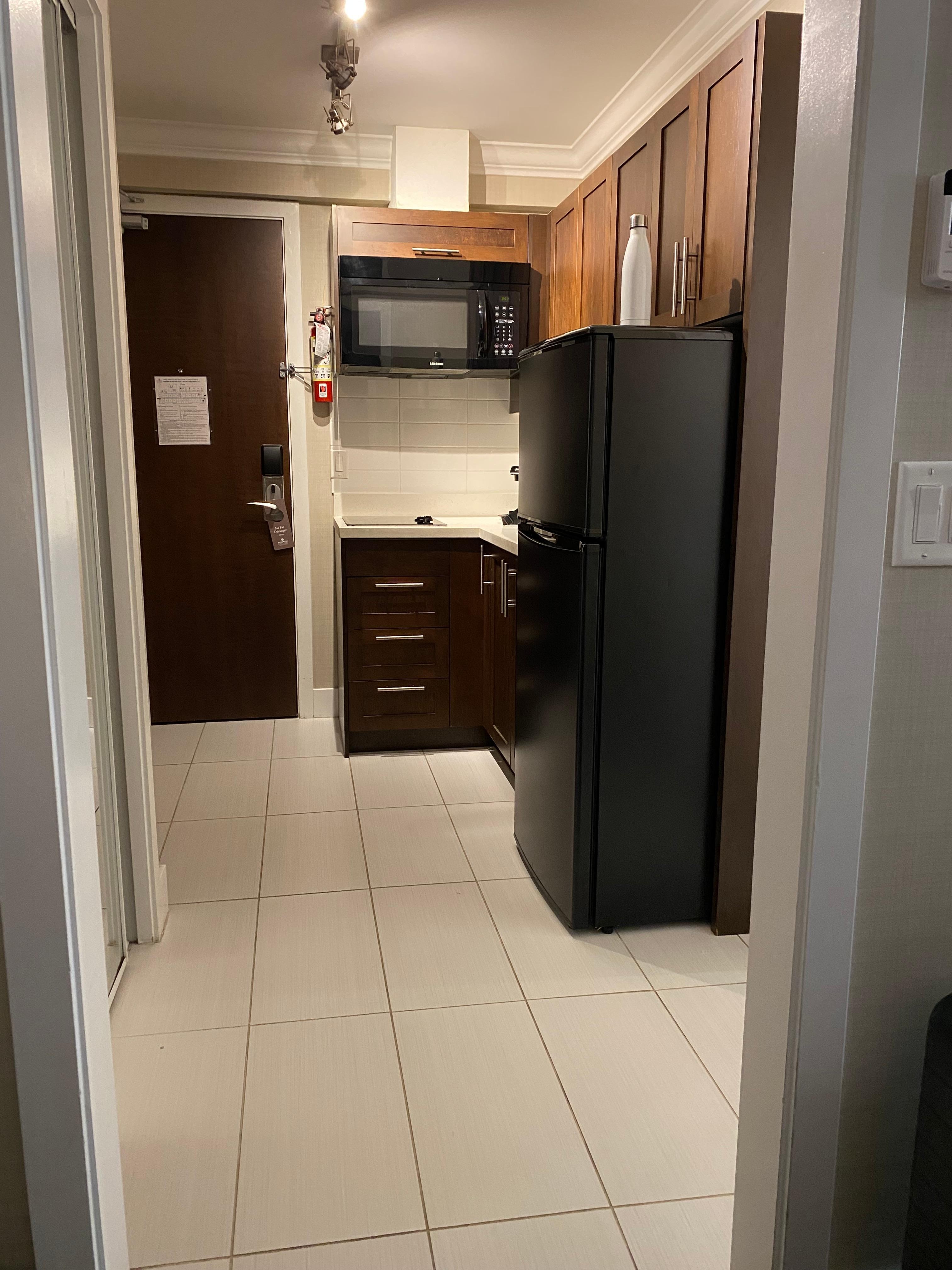 Looking from bedroom doorway to the kitchenette entrance area. Small hot plate style stovetop, full fridge, microwave, dishes, cutlery, pots, toaster and kettle provided. 