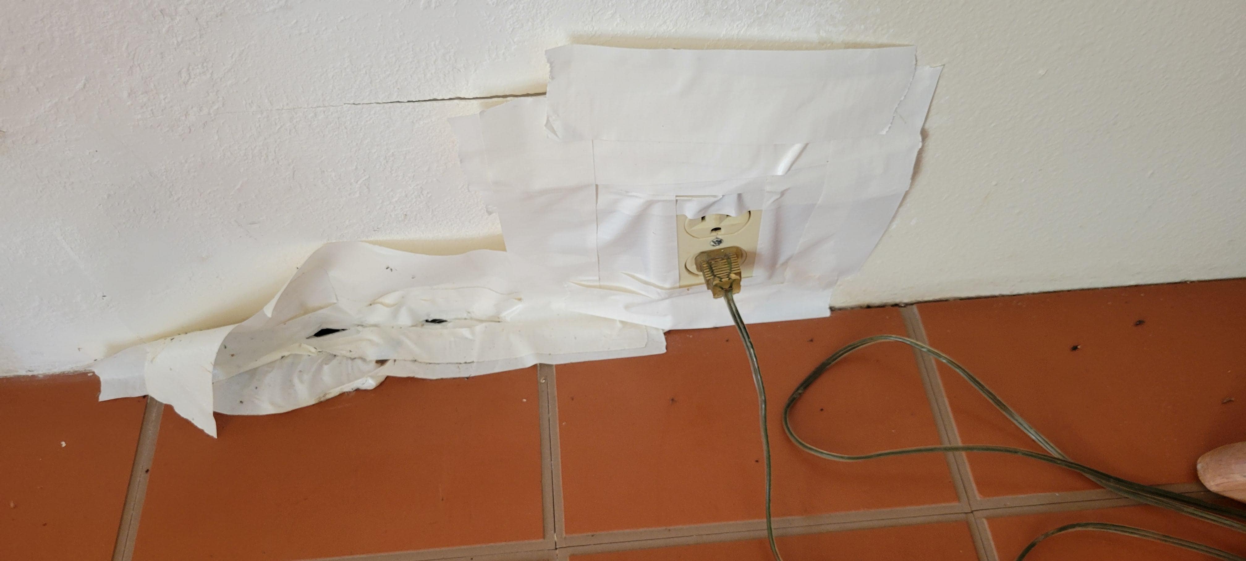 Power plug held in the wall with athletic tape. More tape to the left covering hole in the drywall.