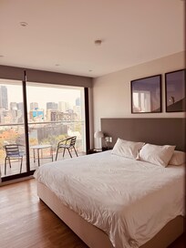 A sleek, sunlit bedroom with minimalist design opens onto a balcony overlooking Mexico City's skyline, blending comfort, style, and urban serenity.