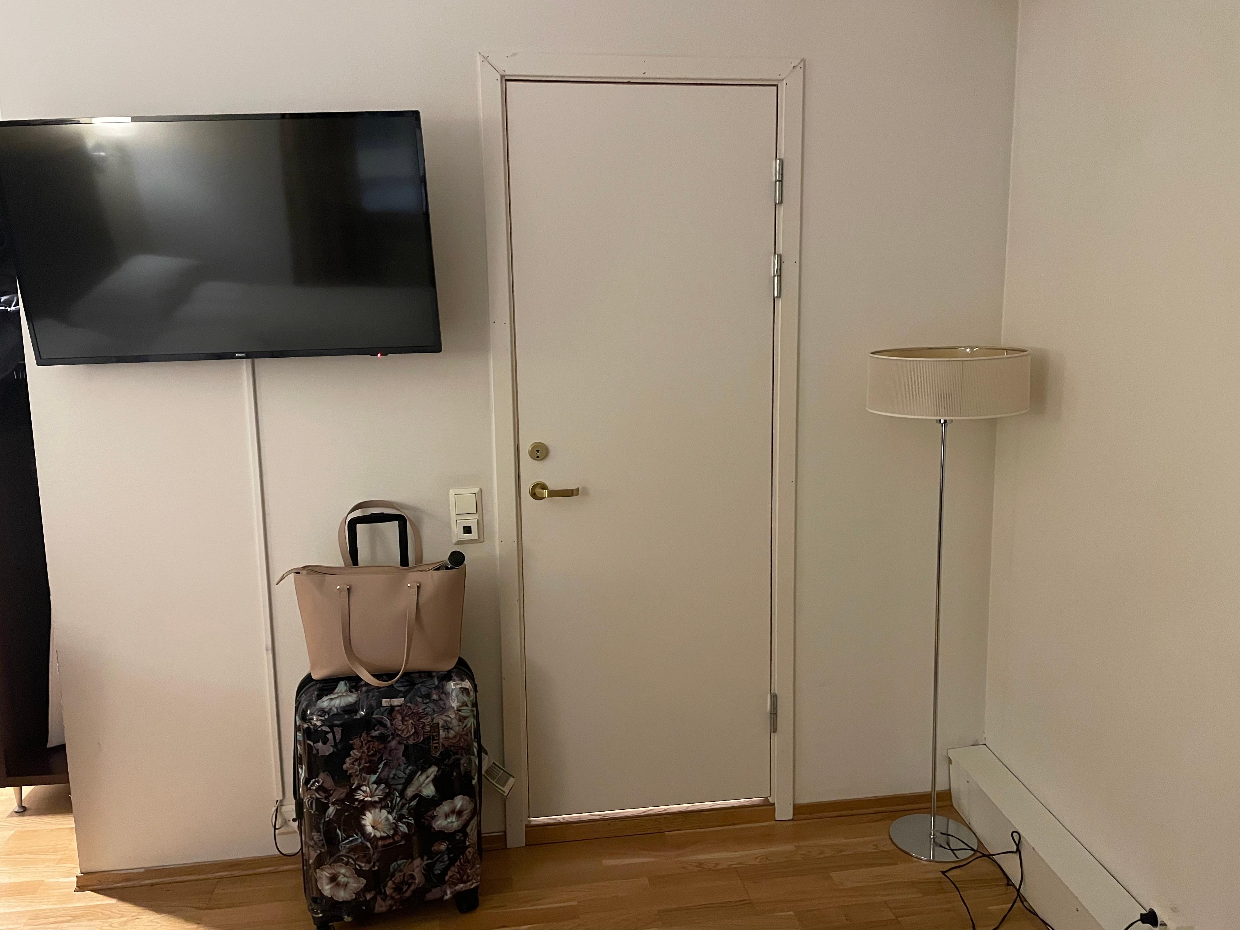 Dør mot bad, tv henger på motsatt vegg av seng. En får ikke frem hvor slitt og skitten stålampen er.