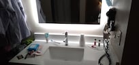Stylish backlit mirror above square Moen sink. Traveler's toiletries are on the counter. To the left, a hanging bar holds clothes. To the right, a mask is hanging, and a hairdryer is plugged into an outlet.