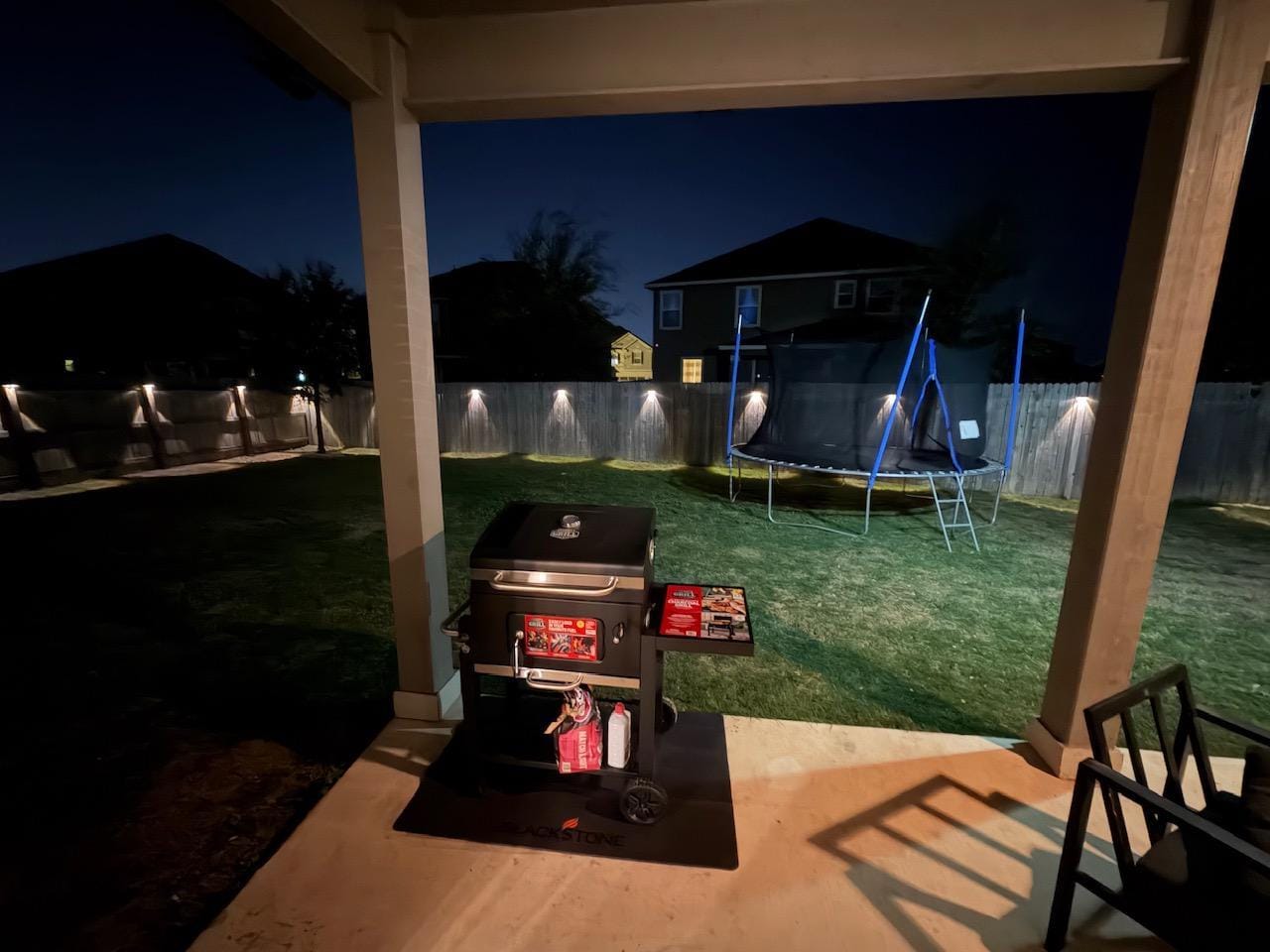 The backyard at night. Beautiful lighting, a trampoline, grill, and cozy little sitting area.