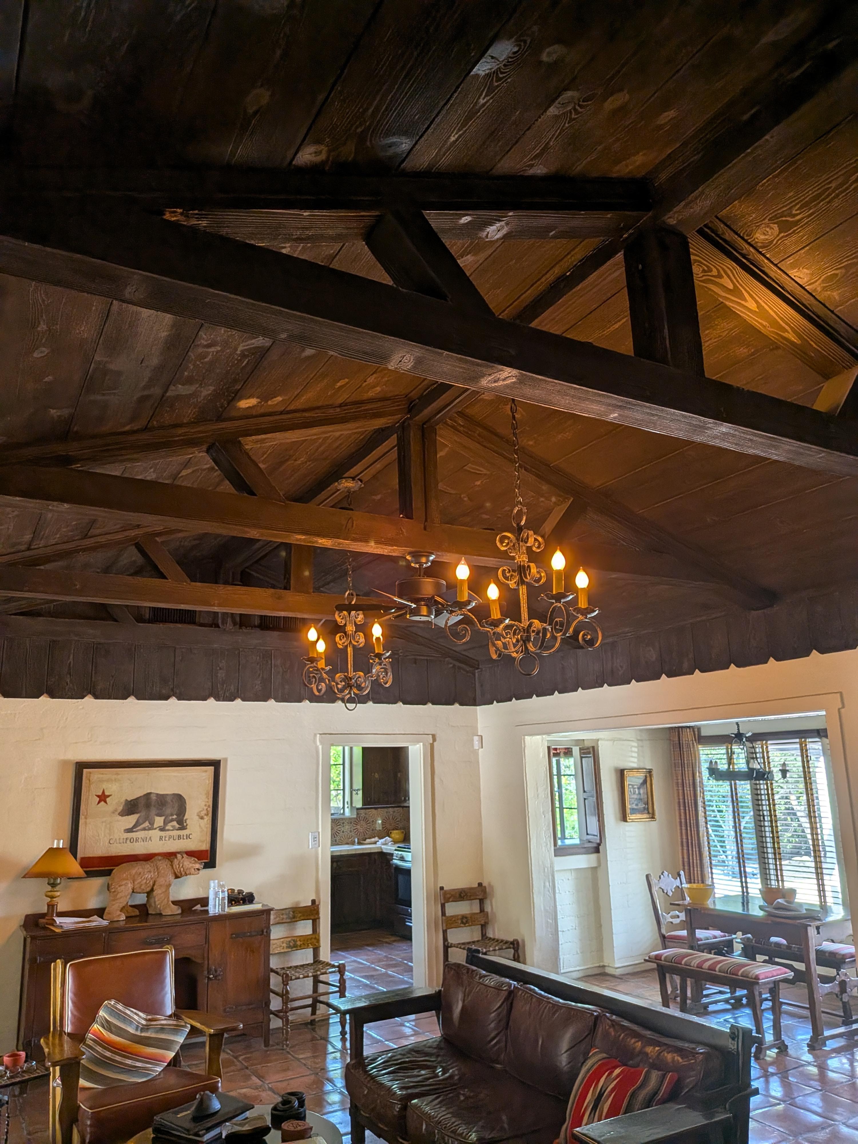 A wide shot of the living and dining area. It features the massive dark wood-vaulted ceilings with original beams, two ornate wrought-iron chandeliers, and the Spanish tile flooring. You can see the California Republic bear art, the leather seating