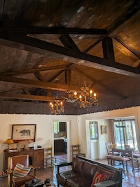 A wide shot of the living and dining area. It features the massive dark wood-vaulted ceilings with original beams, two ornate wrought-iron chandeliers, and the Spanish tile flooring. You can see the California Republic bear art, the leather seating
