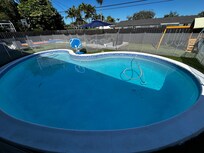 Pool with fence around it & view of the yard activities. There is lounge couch area with umbrella, swing set, mini golf, basketball court, large chess set, ping pong table, & pool basketball hoop.